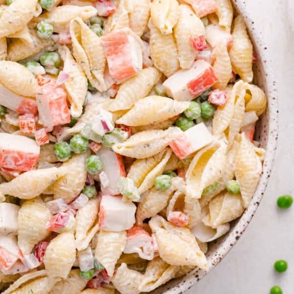 A close-up of a pasta salad with shell pasta, imitation crab meat, green peas, and diced red onion in a creamy dressing.