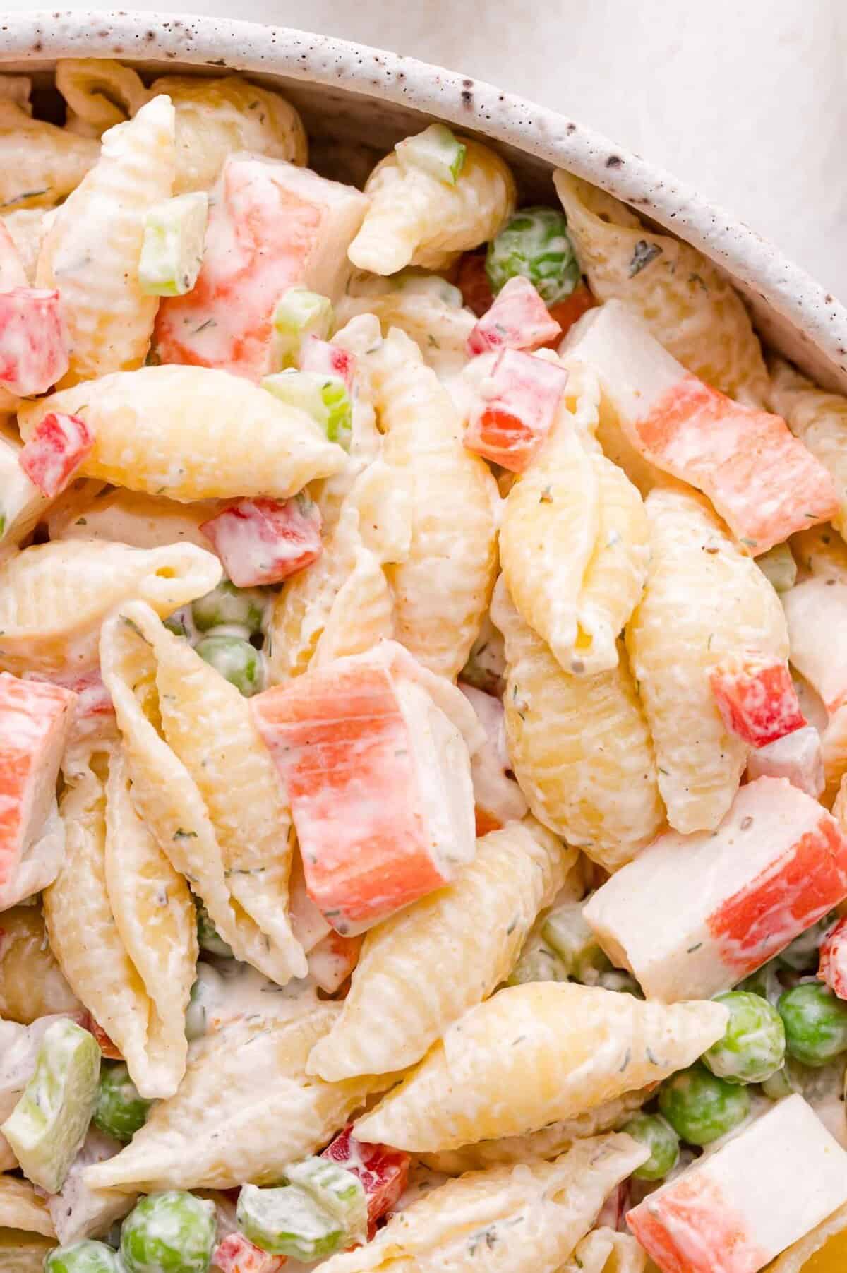 Close-up of pasta shells mixed with imitation crab meat, green peas, diced red bell pepper, and creamy dressing.