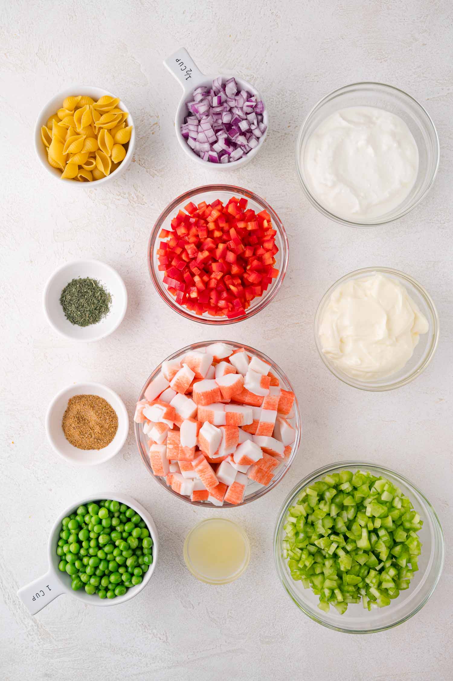 Ingredients for a salad arranged in bowls, including pasta, chopped red onion, bell pepper, peas, celery, imitation crab, seasonings, lemon juice, yogurt, and mayonnaise.