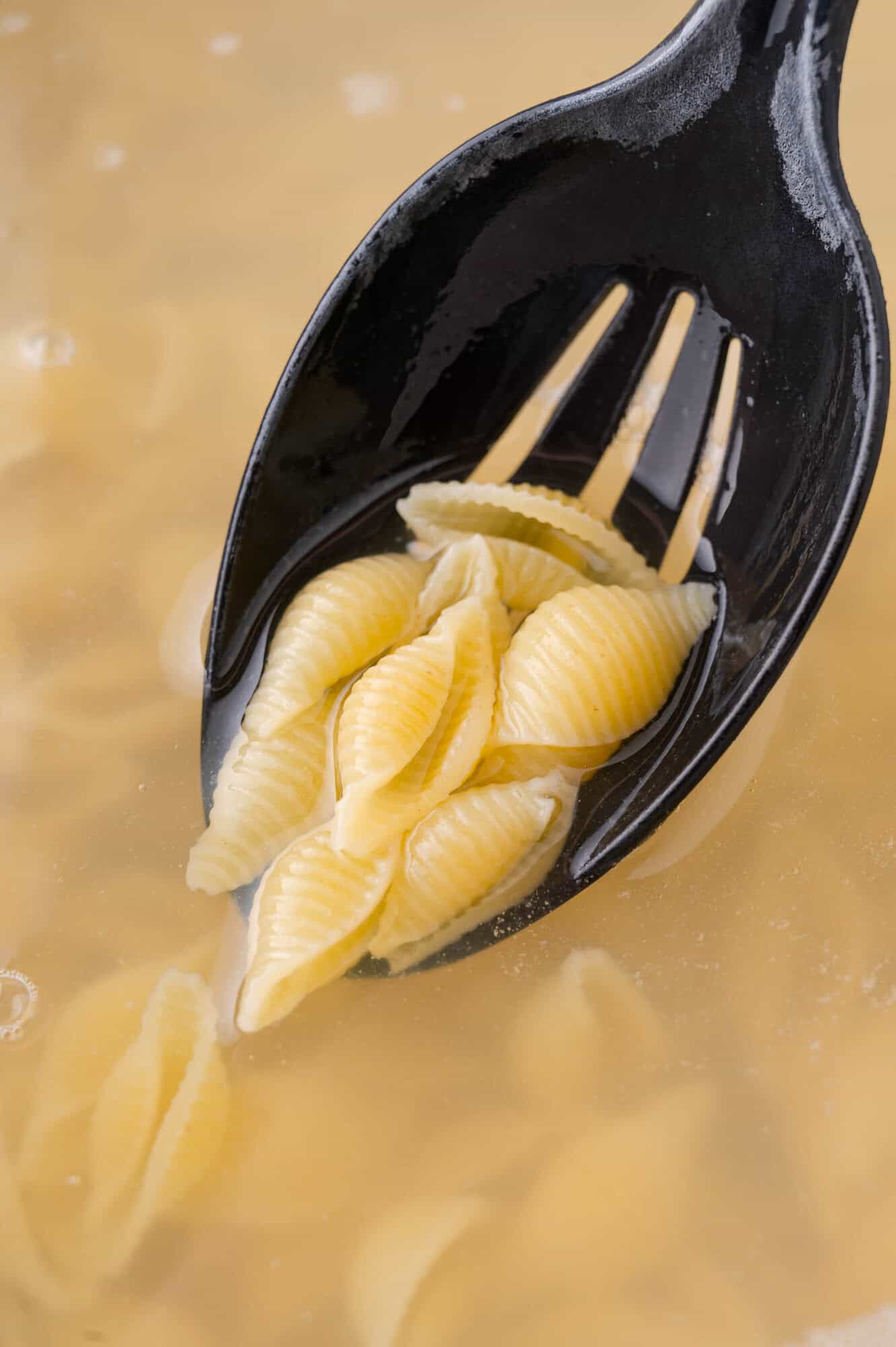A black slotted spoon holding cooked shell pasta above a pot of boiling water.