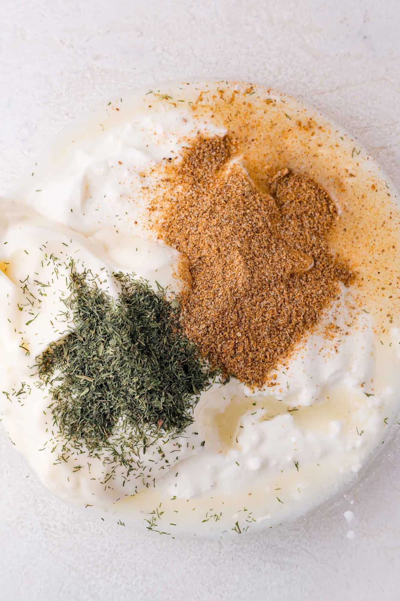A bowl of creamy white sauce topped with dried dill and Old Bay seasoning on a light background.