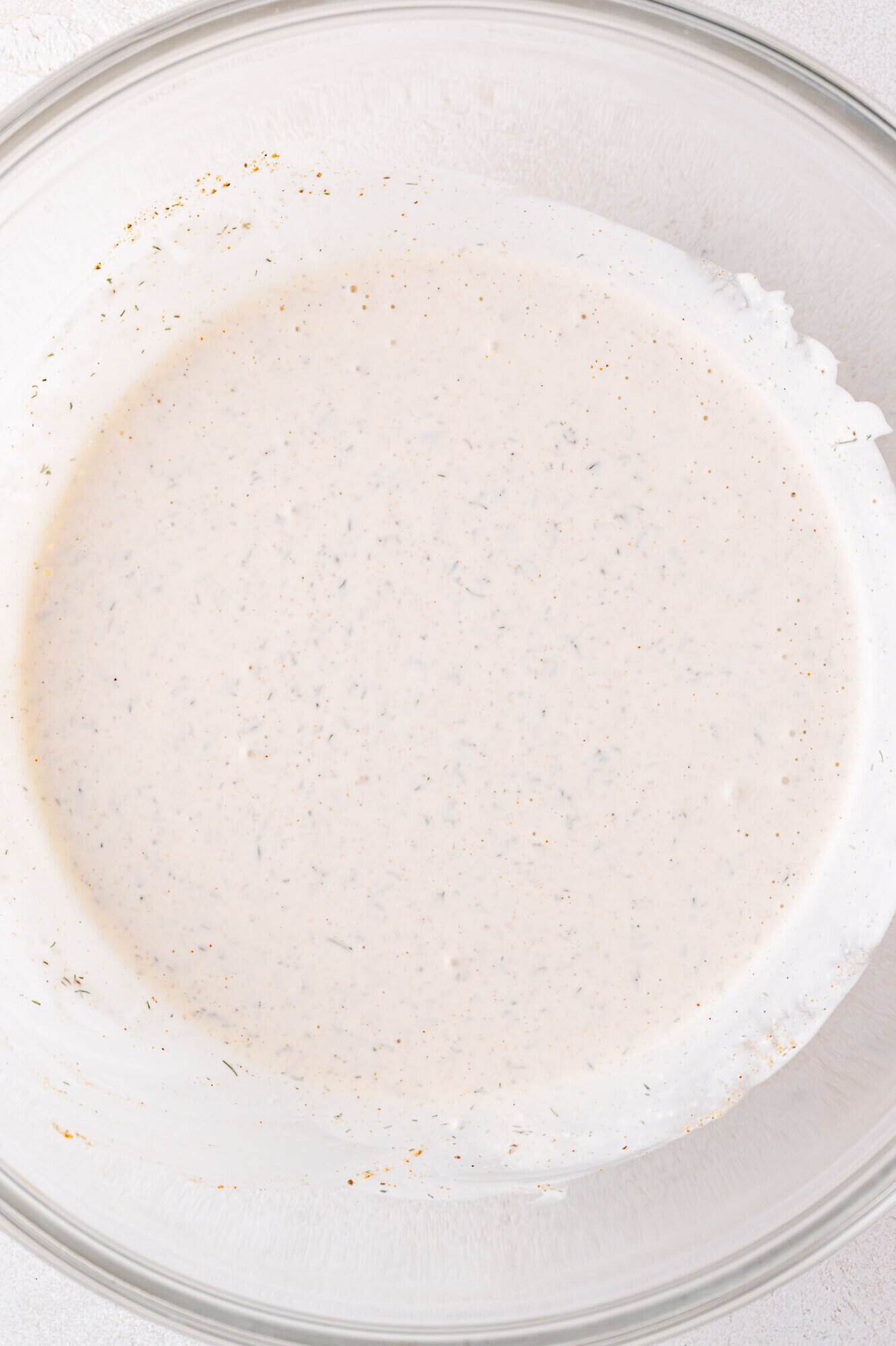 A clear glass bowl filled with a pale, creamy sauce or dressing speckled with herbs and spices.