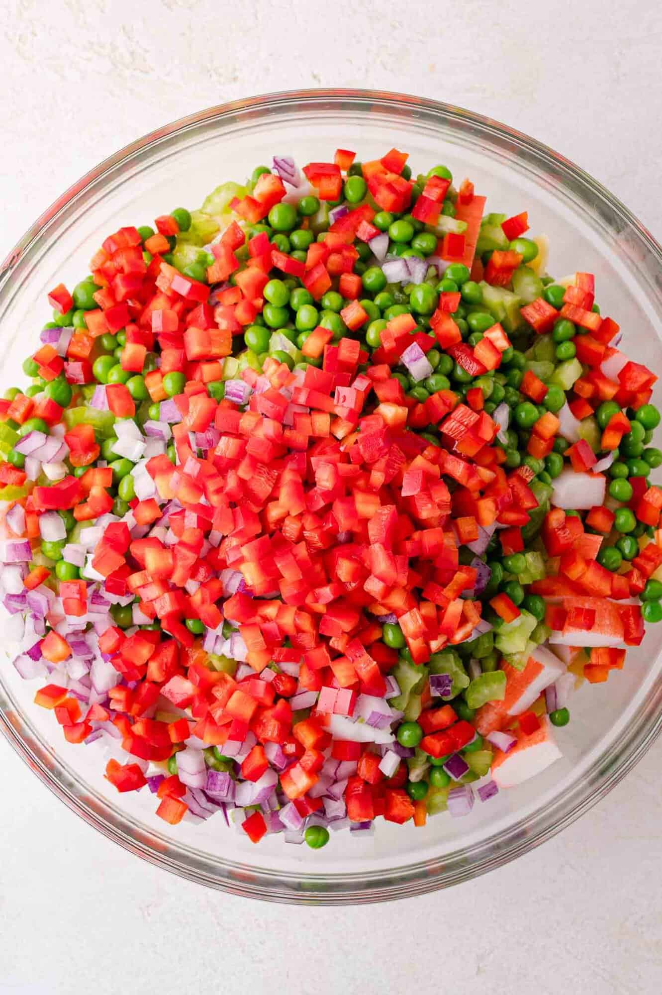 A glass bowl filled with diced red bell peppers, green peas, chopped red onions, and other mixed vegetables.