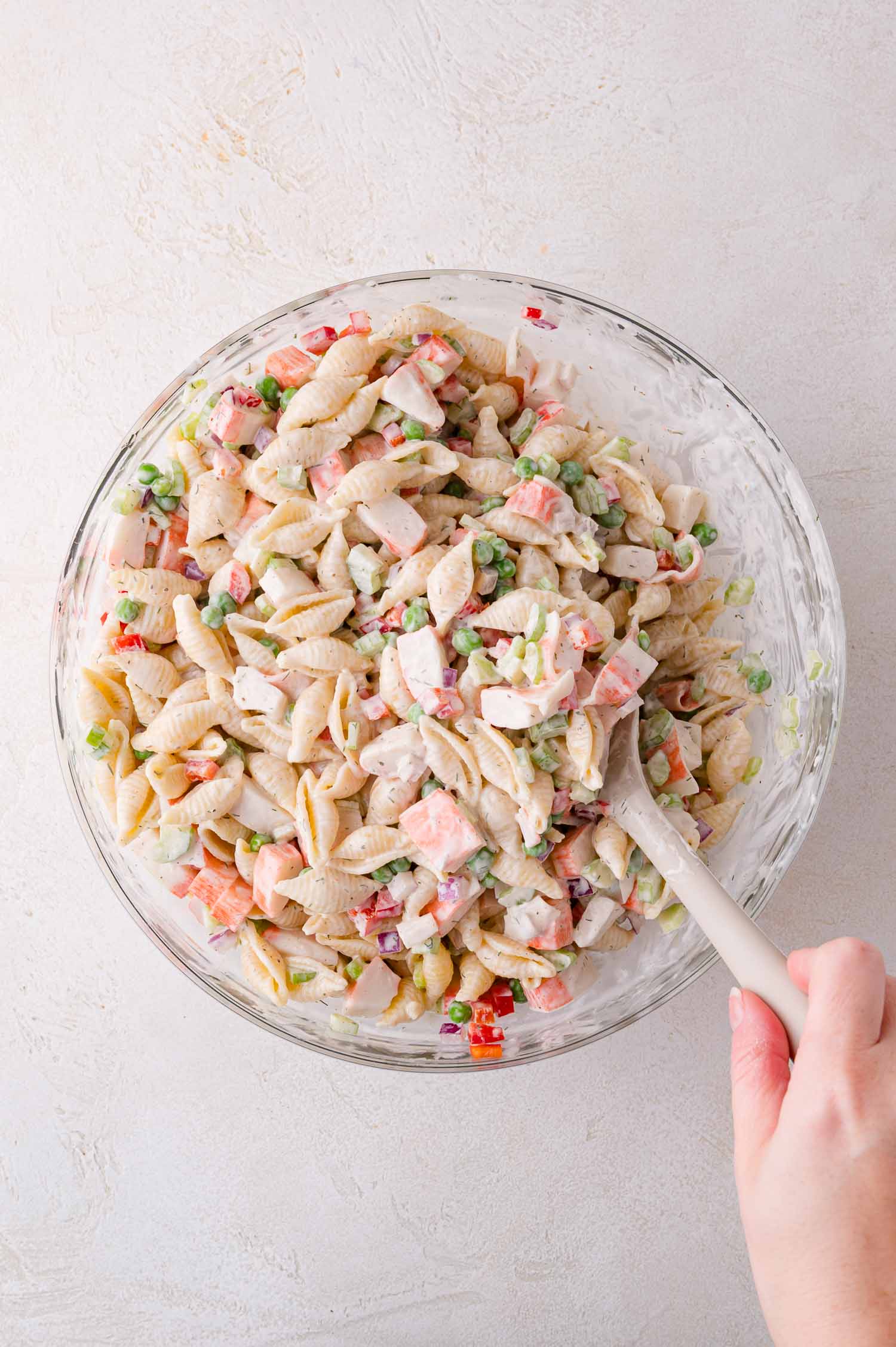 A hand stirs a glass bowl filled with creamy pasta salad containing shell pasta, chopped vegetables, and dressing.