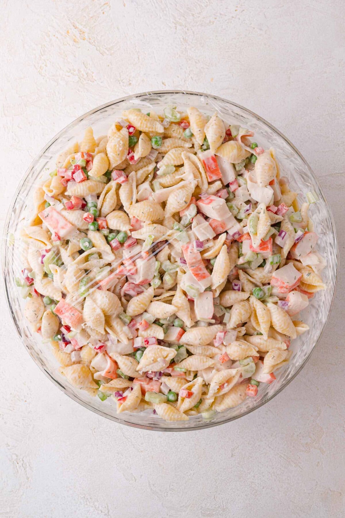 A glass bowl filled with shell pasta salad mixed with imitation crab, chopped vegetables, and a creamy dressing, viewed from above.