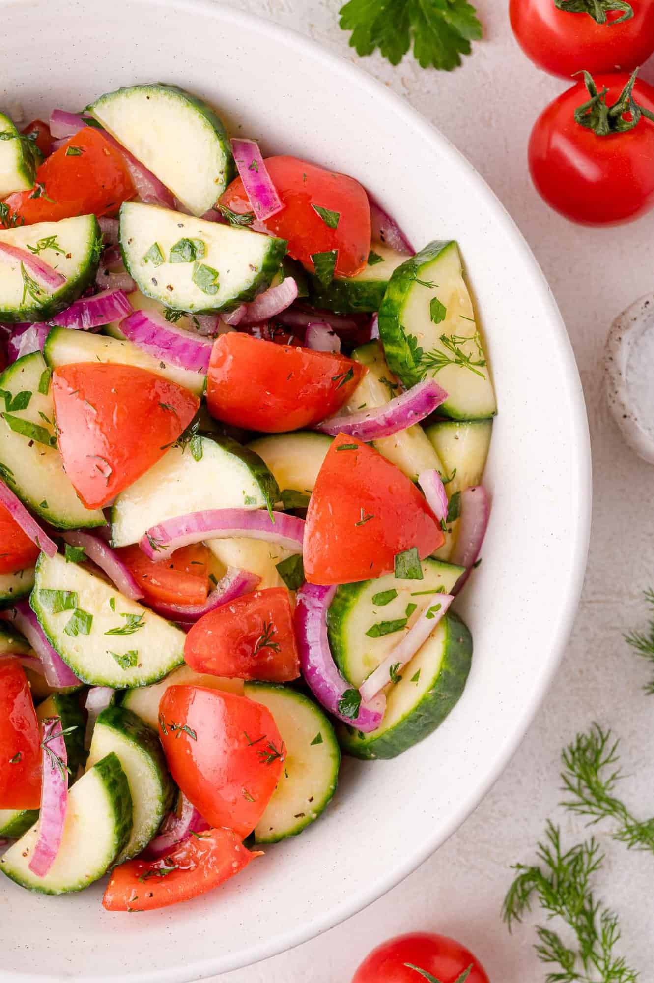 A white bowl filled with salad made of sliced cucumbers, tomatoes, red onions, and herbs, placed on a light surface with fresh tomatoes and herbs nearby.