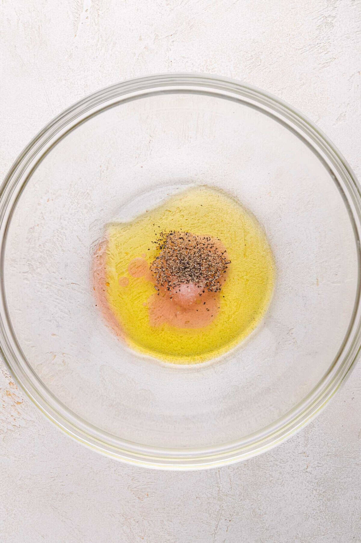 A glass mixing bowl containing olive oil, vinegar, and ground black pepper on a light-colored surface.