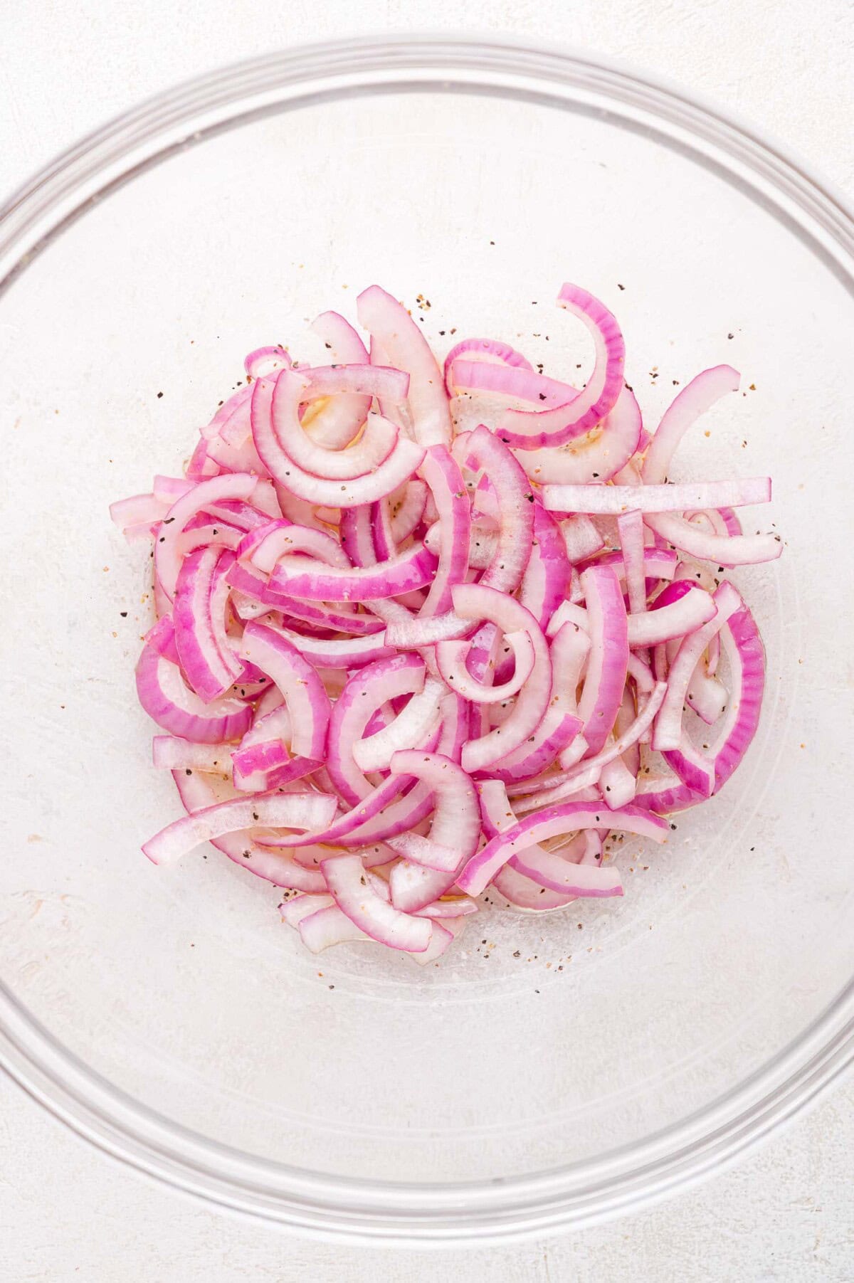 Sliced red onions with black pepper in a clear glass bowl on a light surface.
