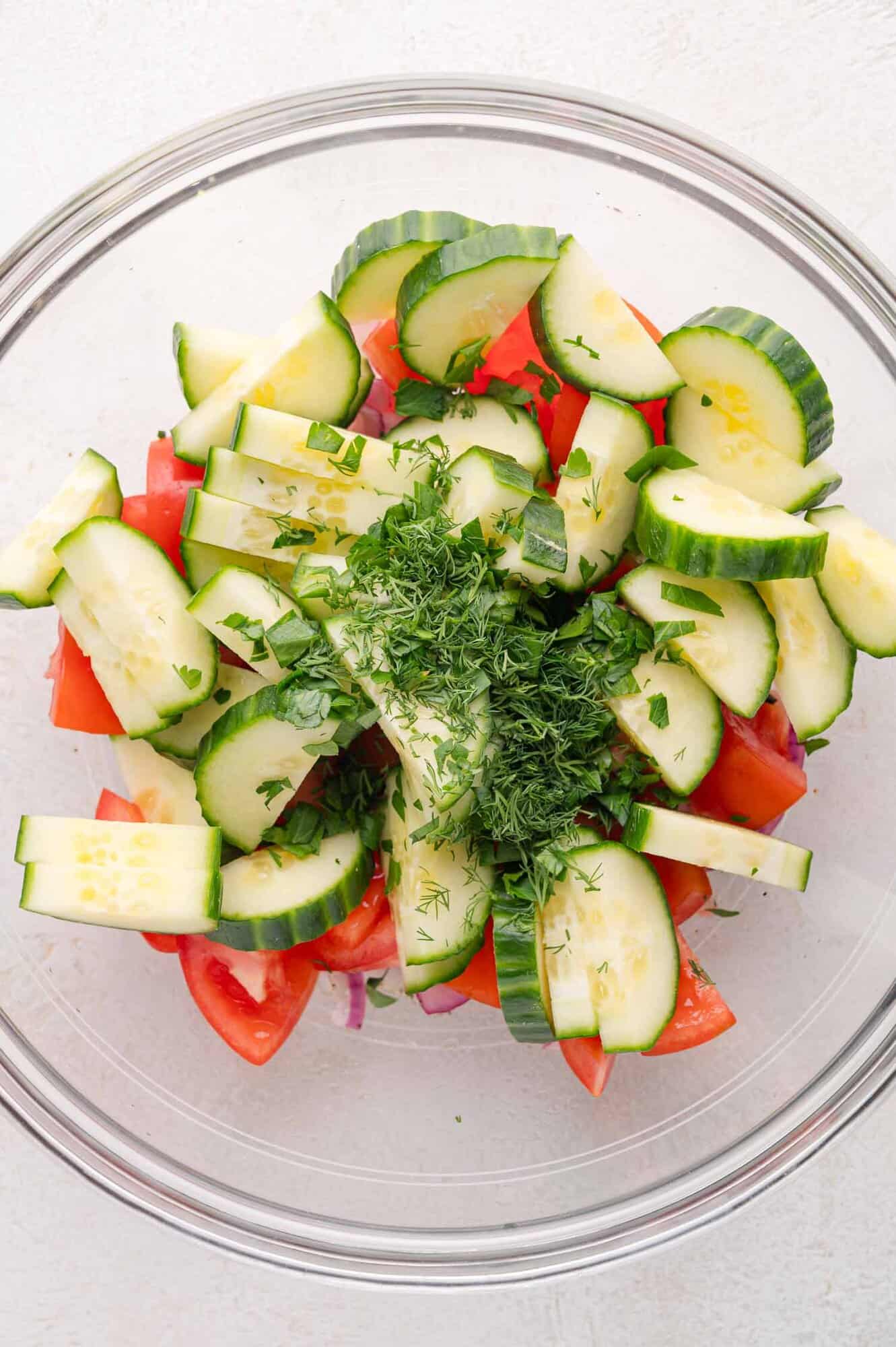 A glass bowl filled with sliced cucumbers, chopped tomatoes, red onions, and fresh chopped herbs on a light surface.