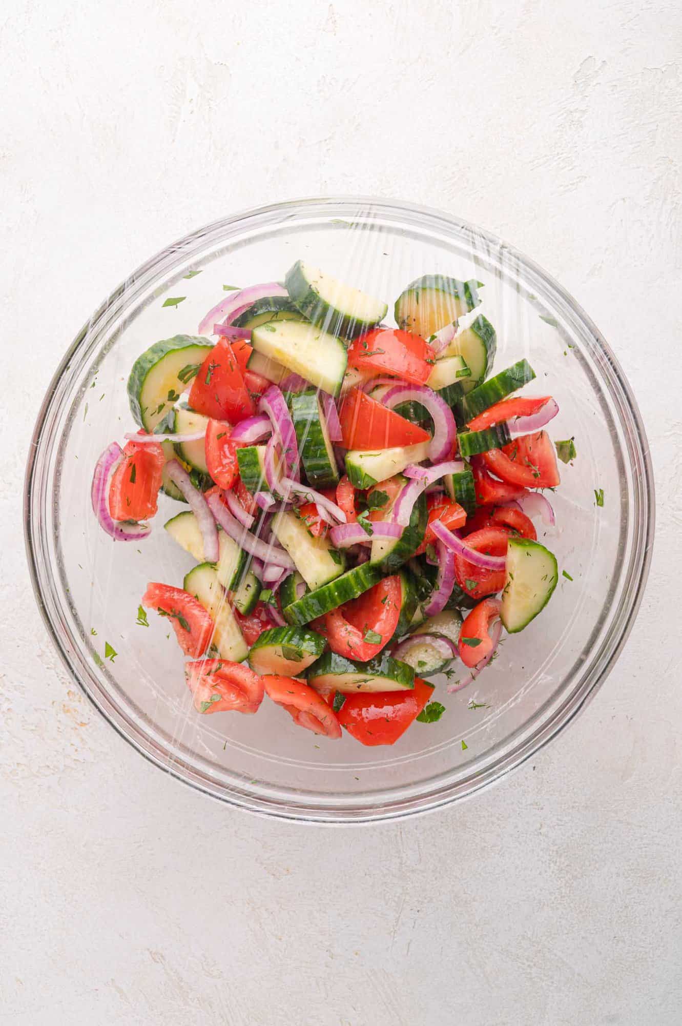 A glass bowl of cucumber, tomato, and red onion salad covered with plastic wrap, viewed from above on a light textured surface.