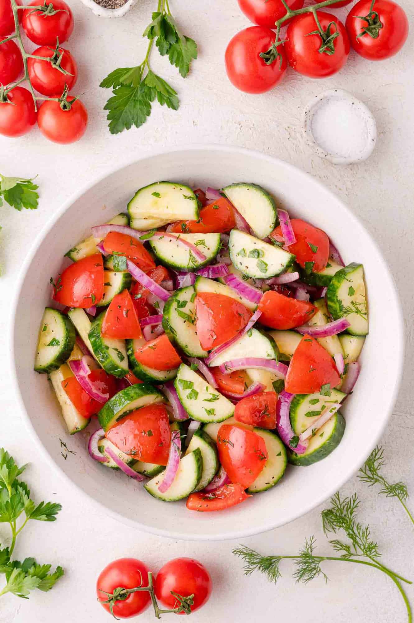 A bowl of cucumber tomato salad garnished with parsley, surrounded by fresh tomatoes, herbs, and a bowl of salt on a white surface.