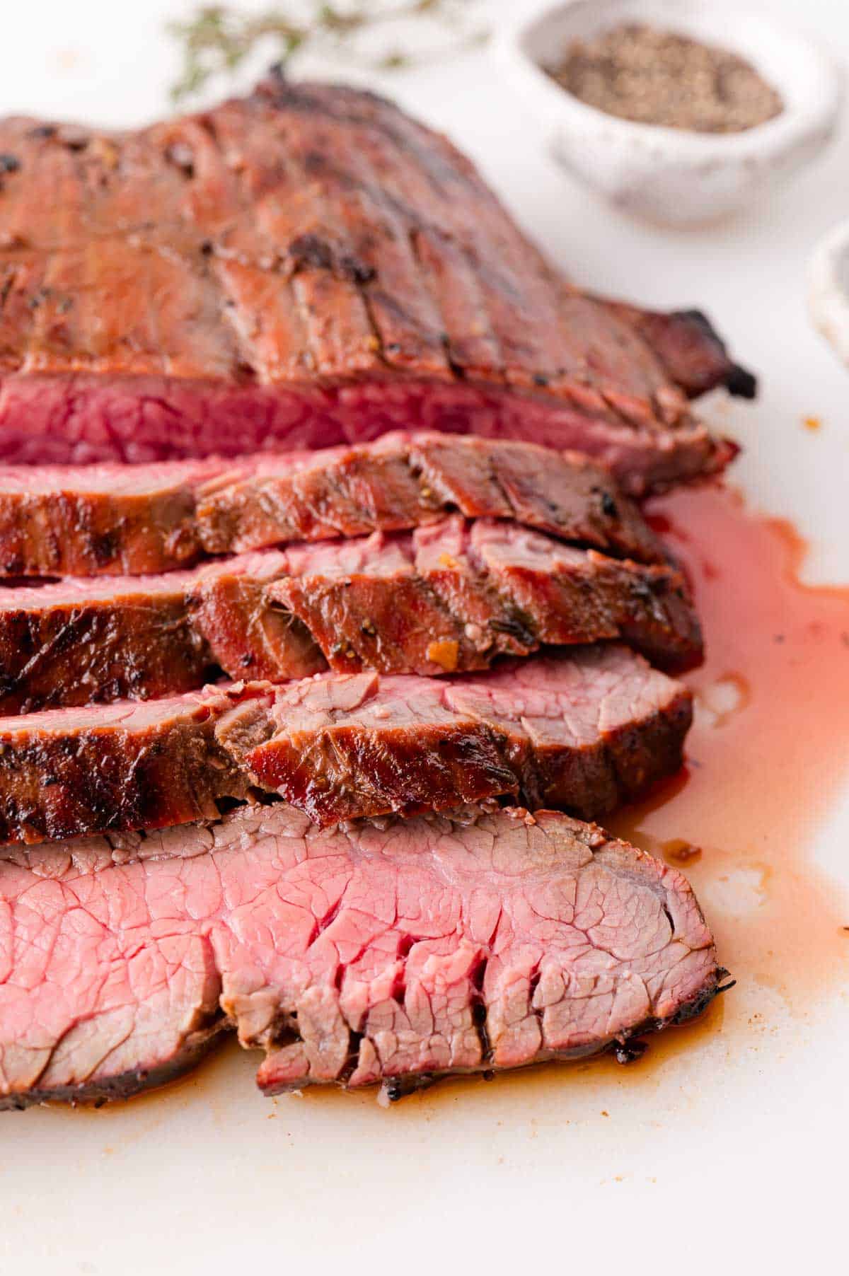 Sliced medium-rare grilled steak on a white surface, with juices visible and a bowl of black pepper in the background.