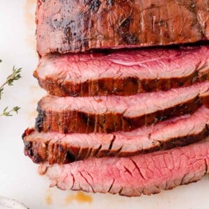 Sliced medium-rare grilled flank steak on a white surface, showing a pink center and seared outer edges. A sprig of thyme is visible to the side.