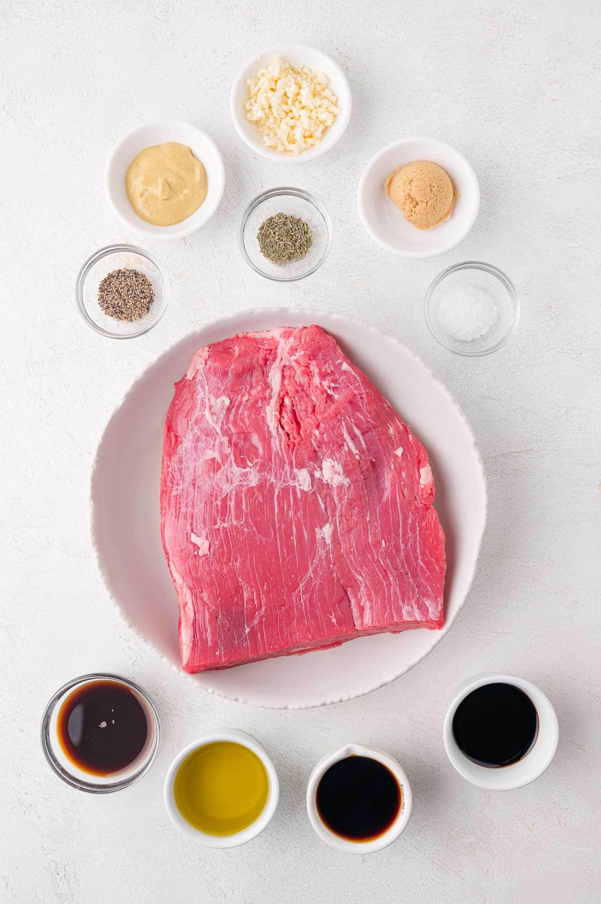 Raw flank steak on a plate surrounded by small bowls containing garlic, mustard, brown sugar, salt, pepper, dried herbs, soy sauce, olive oil, and Worcestershire sauce.