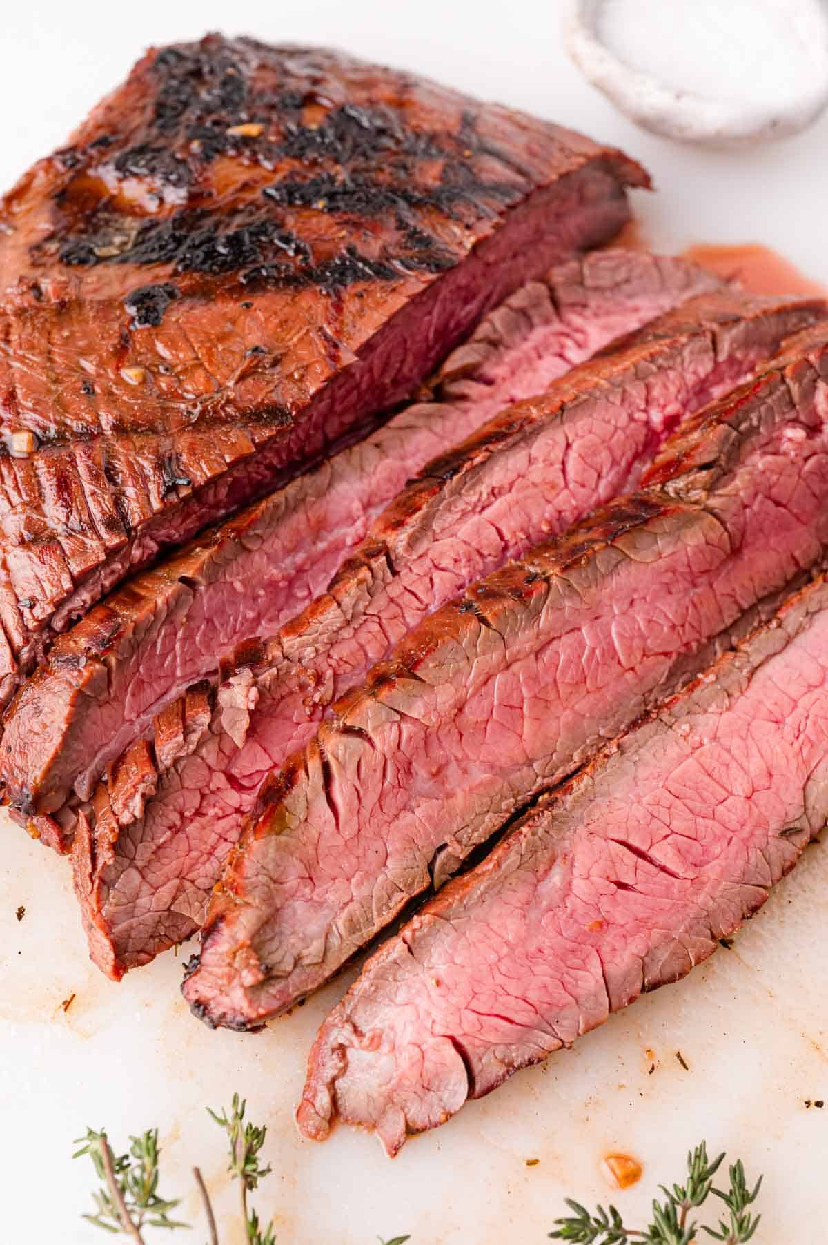 Sliced grilled steak on a cutting board with visible grill marks, served medium rare, with a small dish of salt and sprigs of fresh herbs nearby.