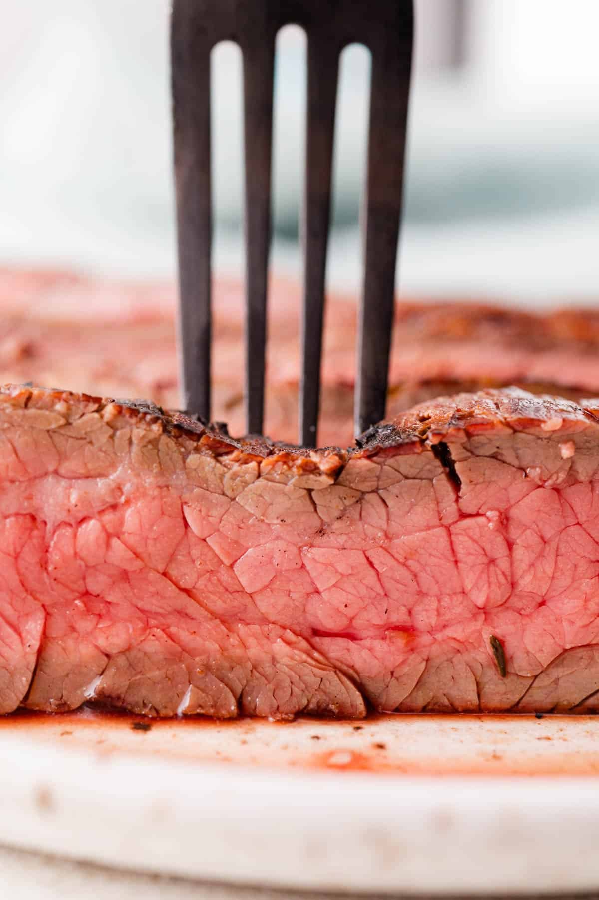 A metal fork pierces a thick slice of rare steak with a seared crust, showcasing its juicy pink interior on a white cutting board.