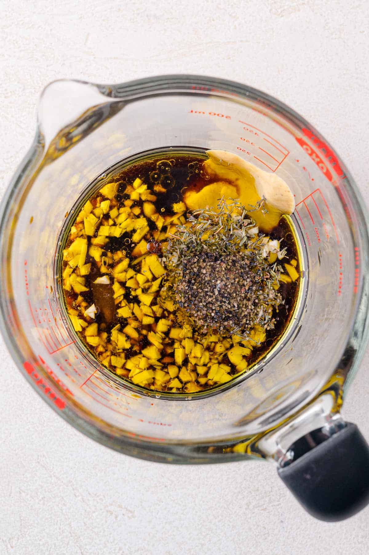 A glass measuring cup filled with chopped garlic, black pepper, dried herbs, soy sauce, oil, and vinegar for making a marinade.