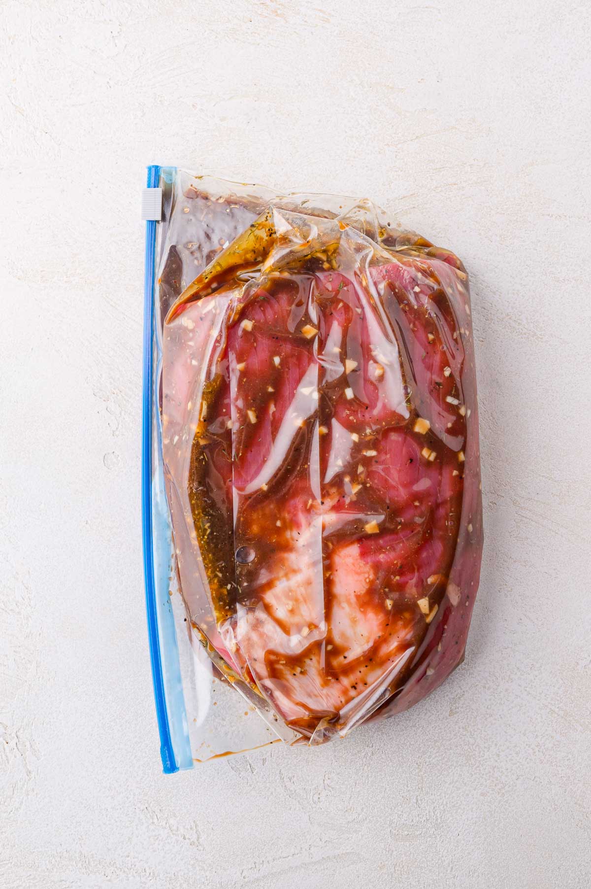 A piece of raw meat marinating in a clear plastic zip-top bag with visible herbs and sauce, placed on a light-colored surface.