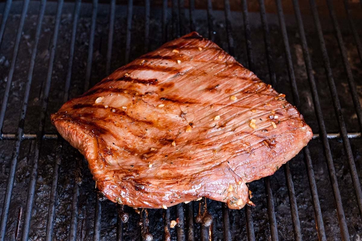 A piece of grilled flank steak with sear marks is cooking on a barbecue grill.
