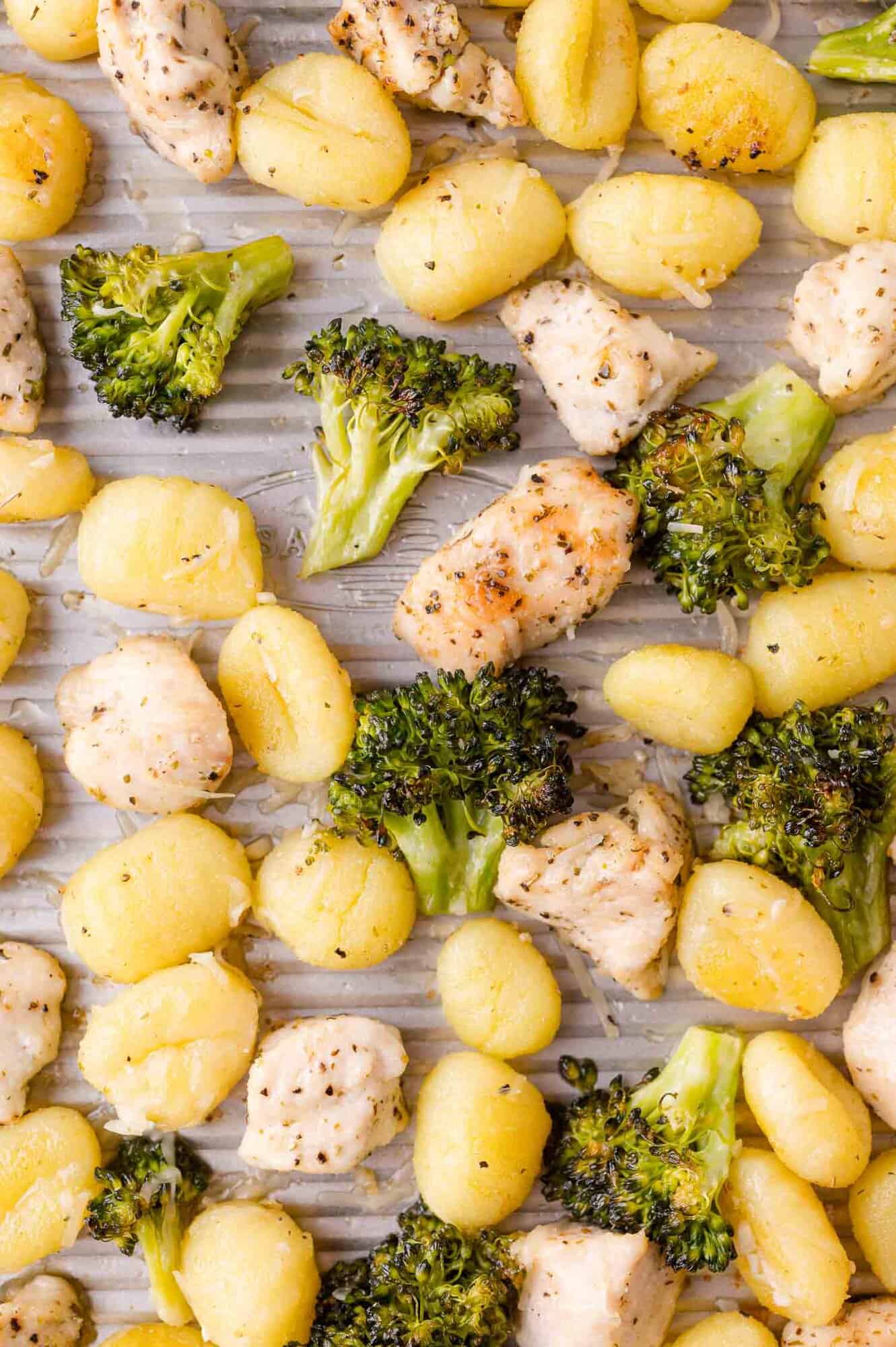 Garlic butter gnocchi, broccoli florets, and seasoned chicken pieces spread out on a lined baking sheet.