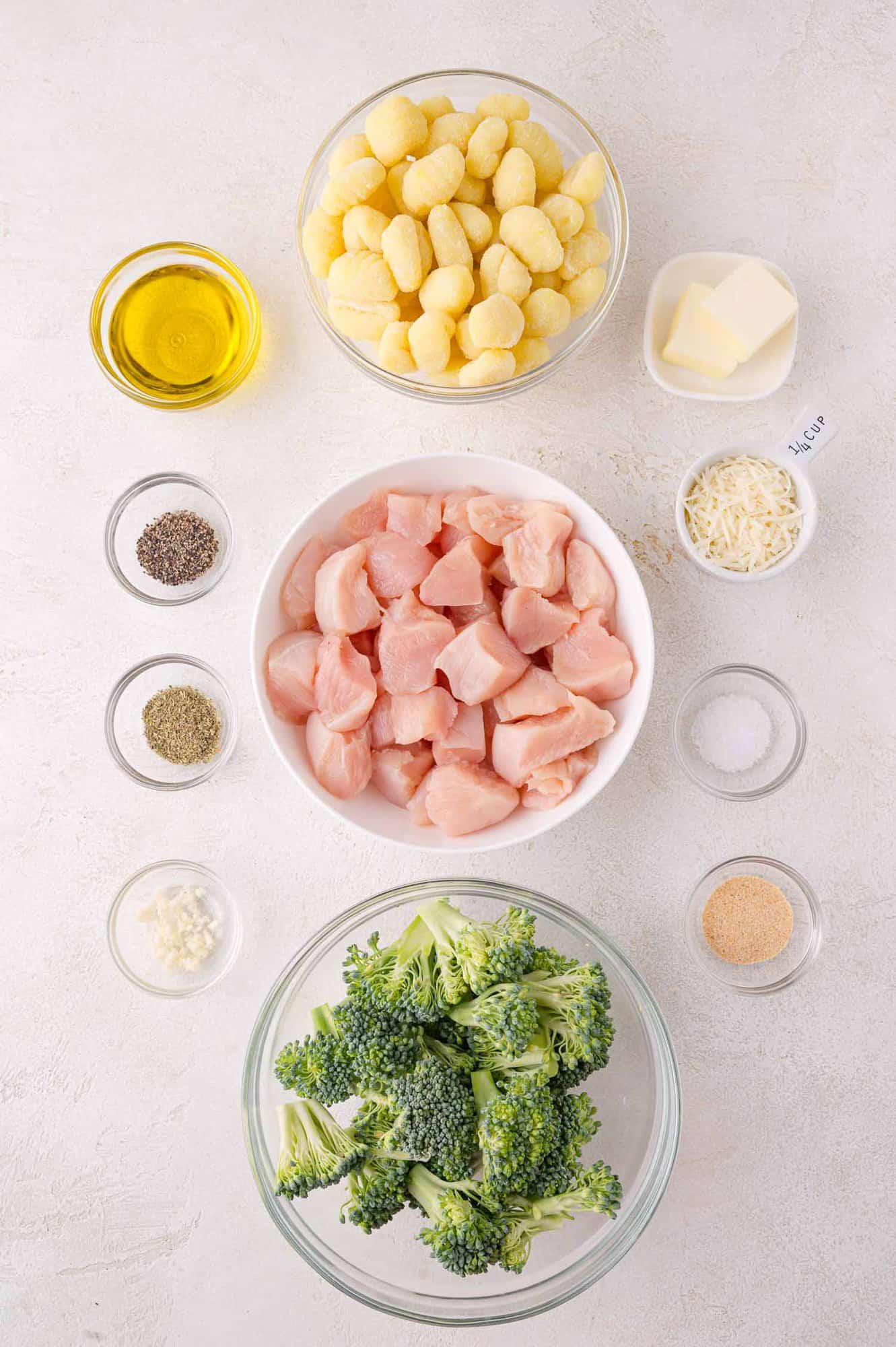 Top-down view of raw ingredients in bowls: gnocchi, chicken breast pieces, broccoli florets, olive oil, butter, grated cheese, minced garlic, and seasonings on a white surface.