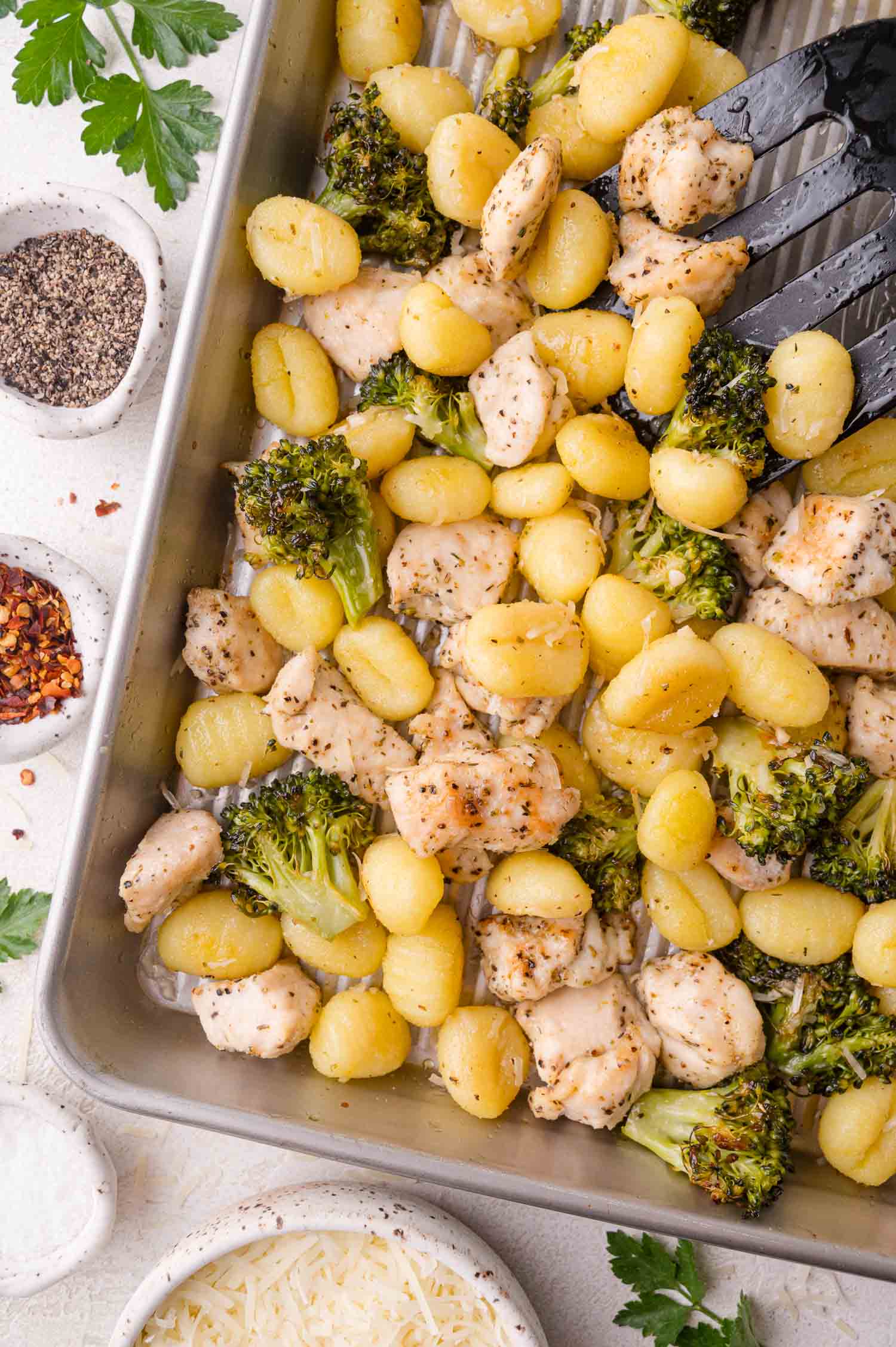 Baked gnocchi with broccoli and chicken pieces in a metal dish, surrounded by bowls of grated cheese, pepper, and red pepper flakes.