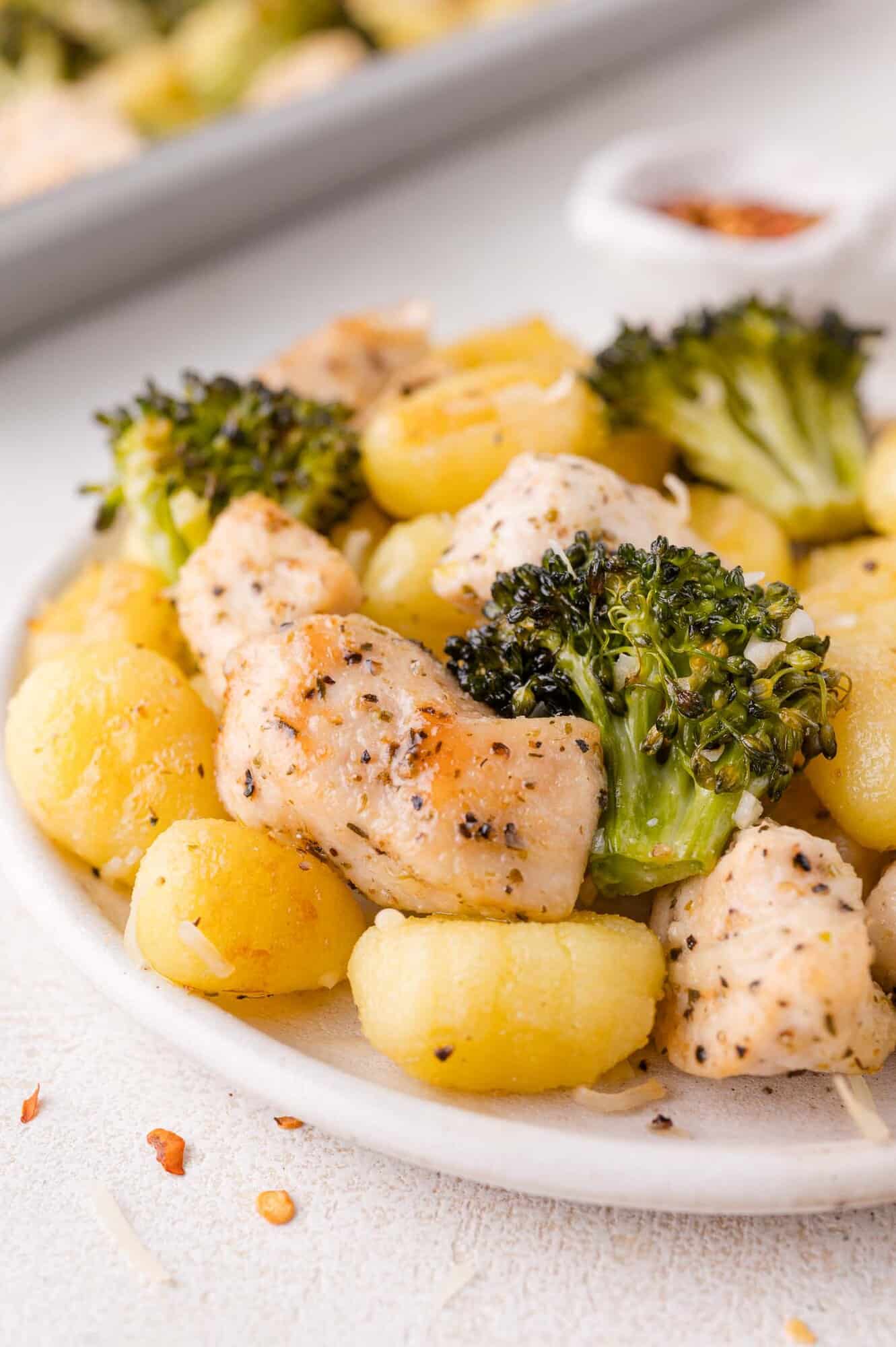 A plate of cooked gnocchi, seasoned chicken pieces, and broccoli florets, garnished with spices and grated cheese.