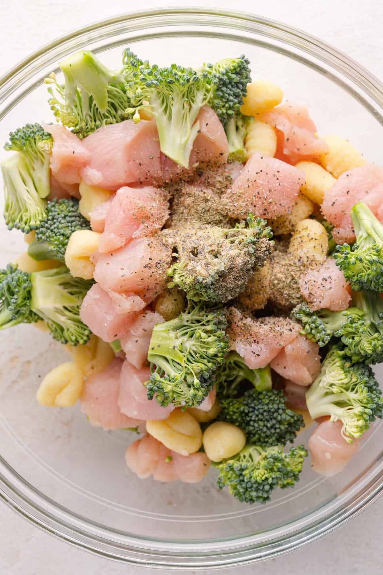 A glass bowl filled with raw diced chicken, broccoli florets, gnocchi, and mixed spices.