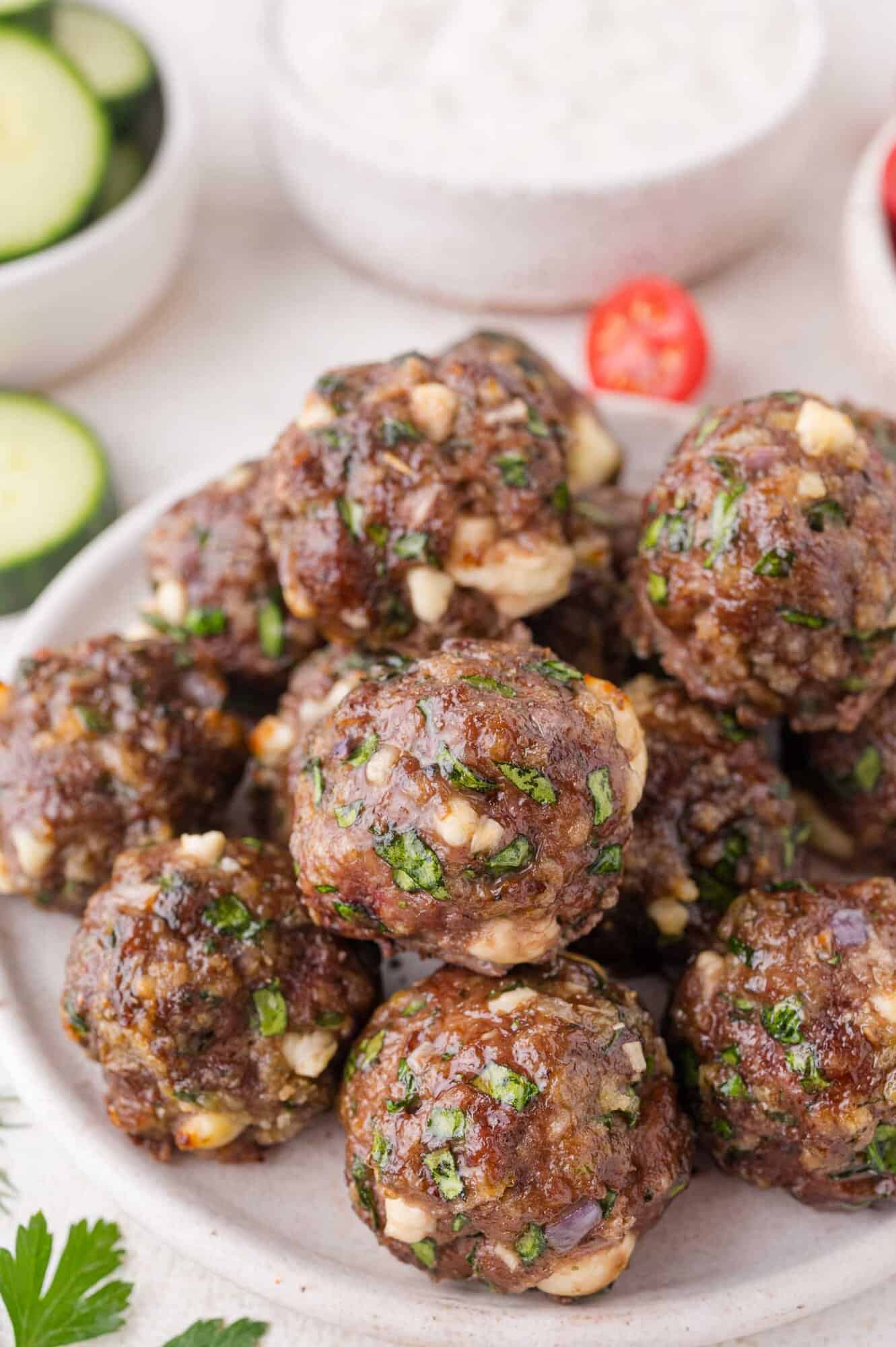 A plate of cooked Greek meatballs mixed with spinach and cheese, with bowls of sliced cucumber and sauce in the background.