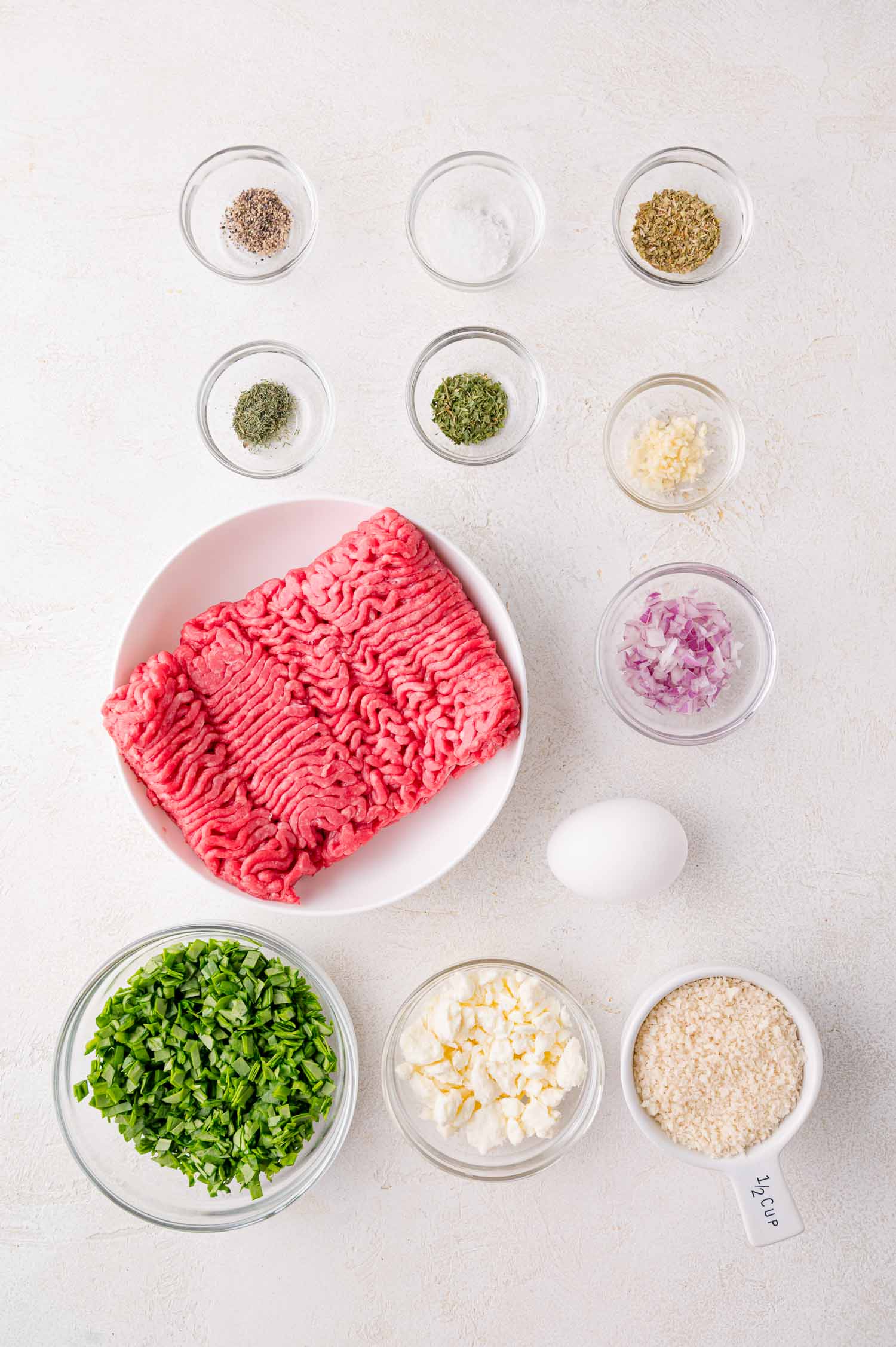 Raw ground meat, chopped spinach, diced onion, minced garlic, feta cheese, egg, breadcrumbs, and assorted seasonings are arranged in separate bowls on a white surface.