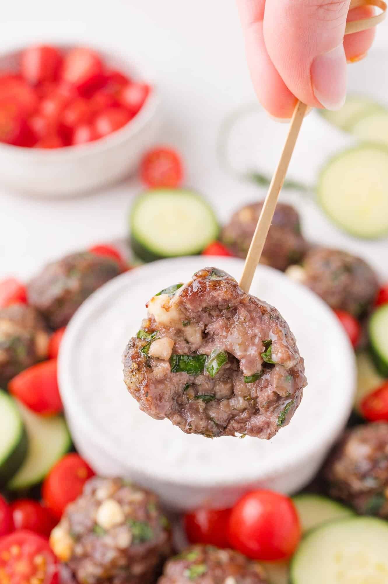 A hand holds a Greek meatball on a skewer above a bowl of white dipping sauce, surrounded by sliced cucumbers and cherry tomatoes.
