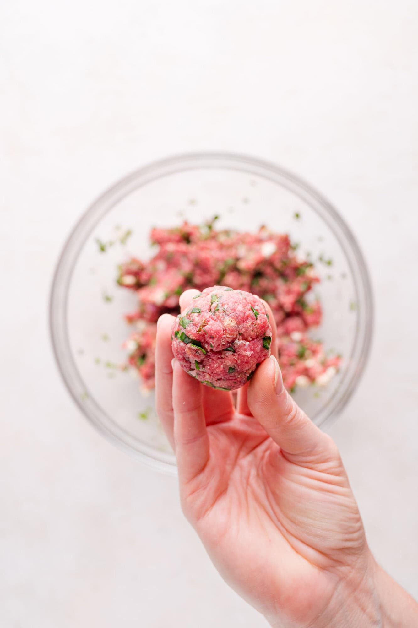 A hand holds a raw meatball in front of a glass bowl filled with ground meat mixture.