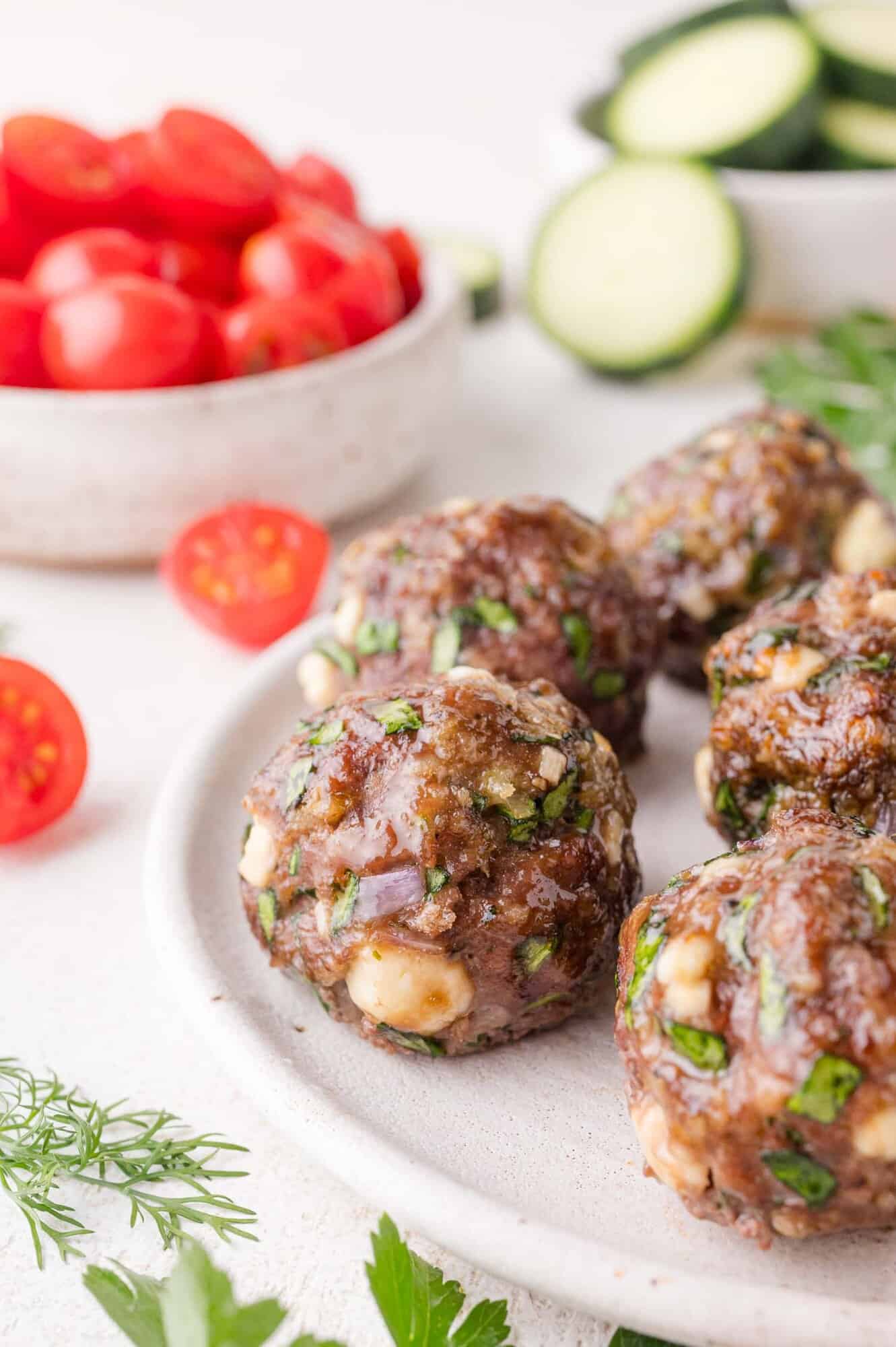 Four cooked Greek meatballs with herbs and cheese on a plate, with sliced cherry tomatoes and cucumber in the background.