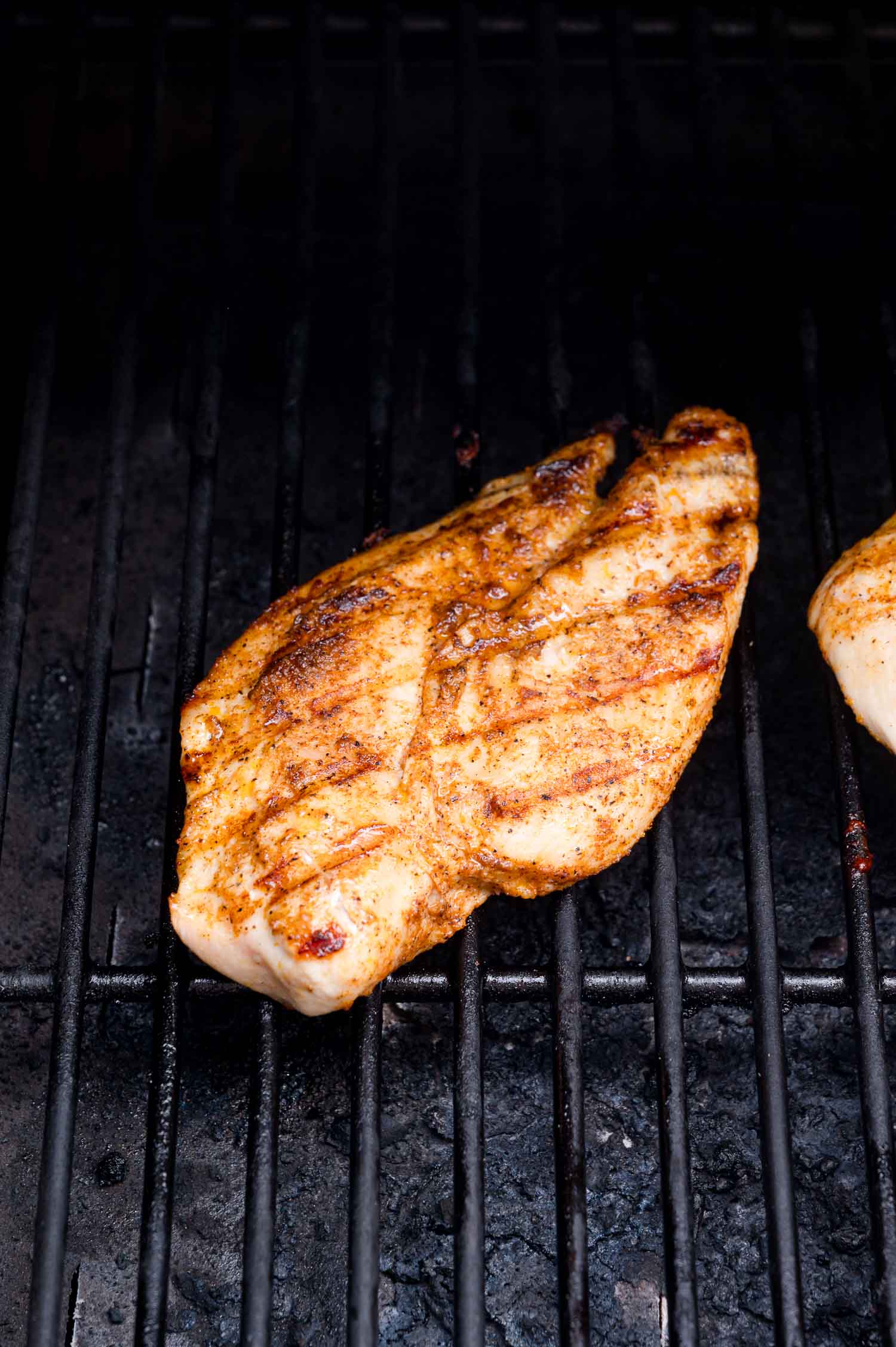 A seasoned chicken breast is cooking on a grill with visible grill marks and char on the surface.