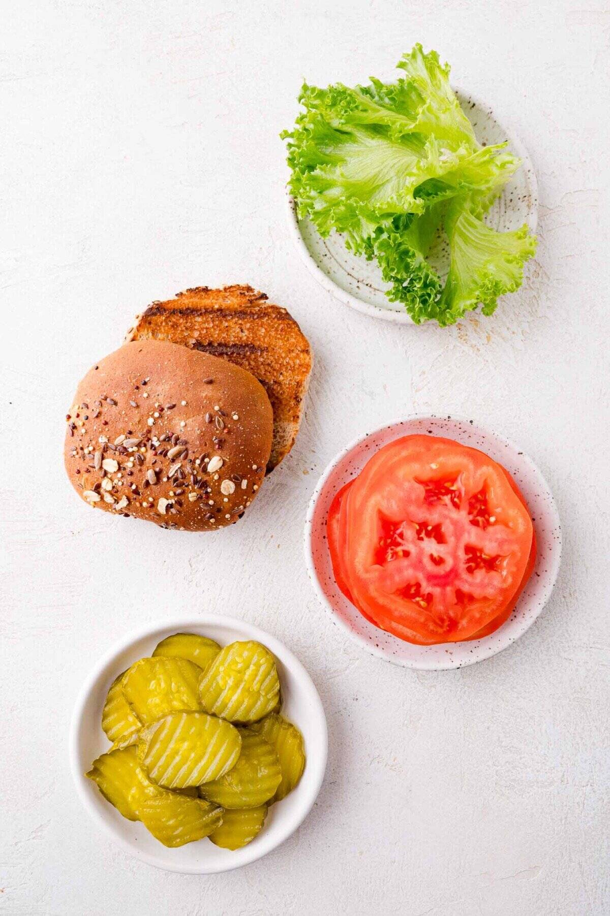 A plate with green lettuce, a sliced wheat burger bun, a bowl of pickle slices, and a plate with two tomato slices arranged on a white surface.