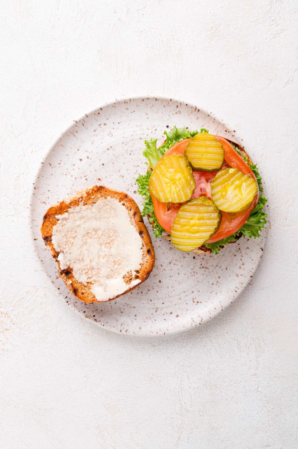 An open-faced sandwich on a speckled plate with lettuce, tomato, pickles, and mayonnaise on a sesame seed bun.
