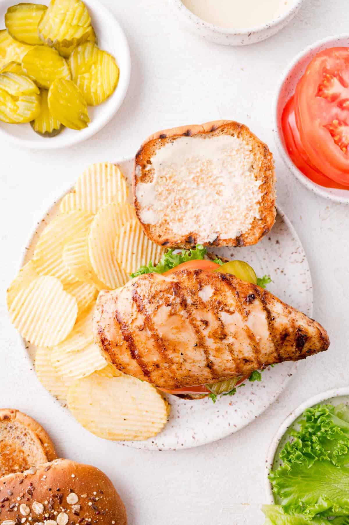 Grilled chicken sandwich with lettuce on a bun, served with ridged potato chips, sliced pickles, tomato slices, and a bowl of sauce on a white surface.
