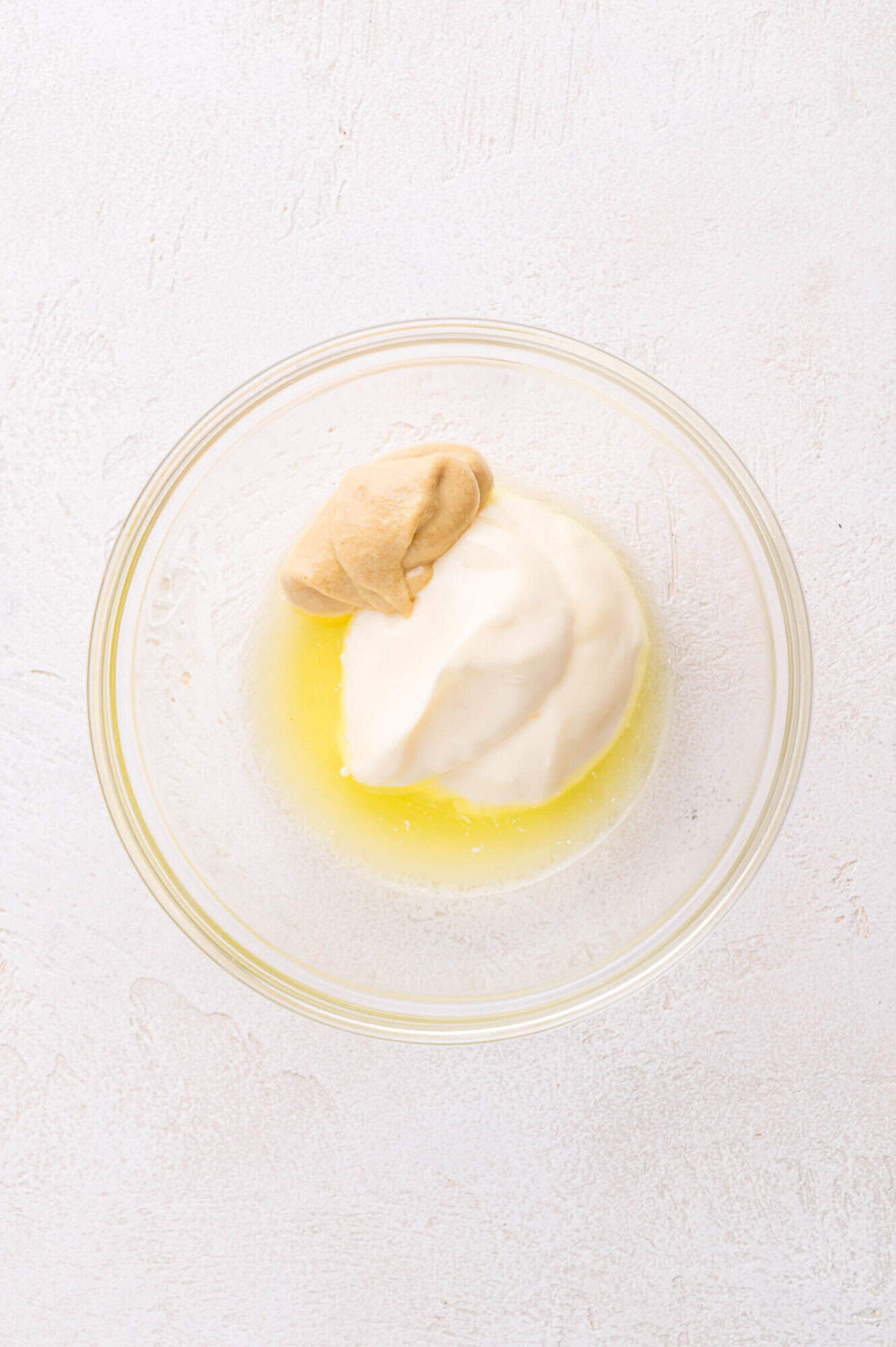 A glass bowl containing mayonnaise, yellow mustard, and pickle juice on a light-colored surface.