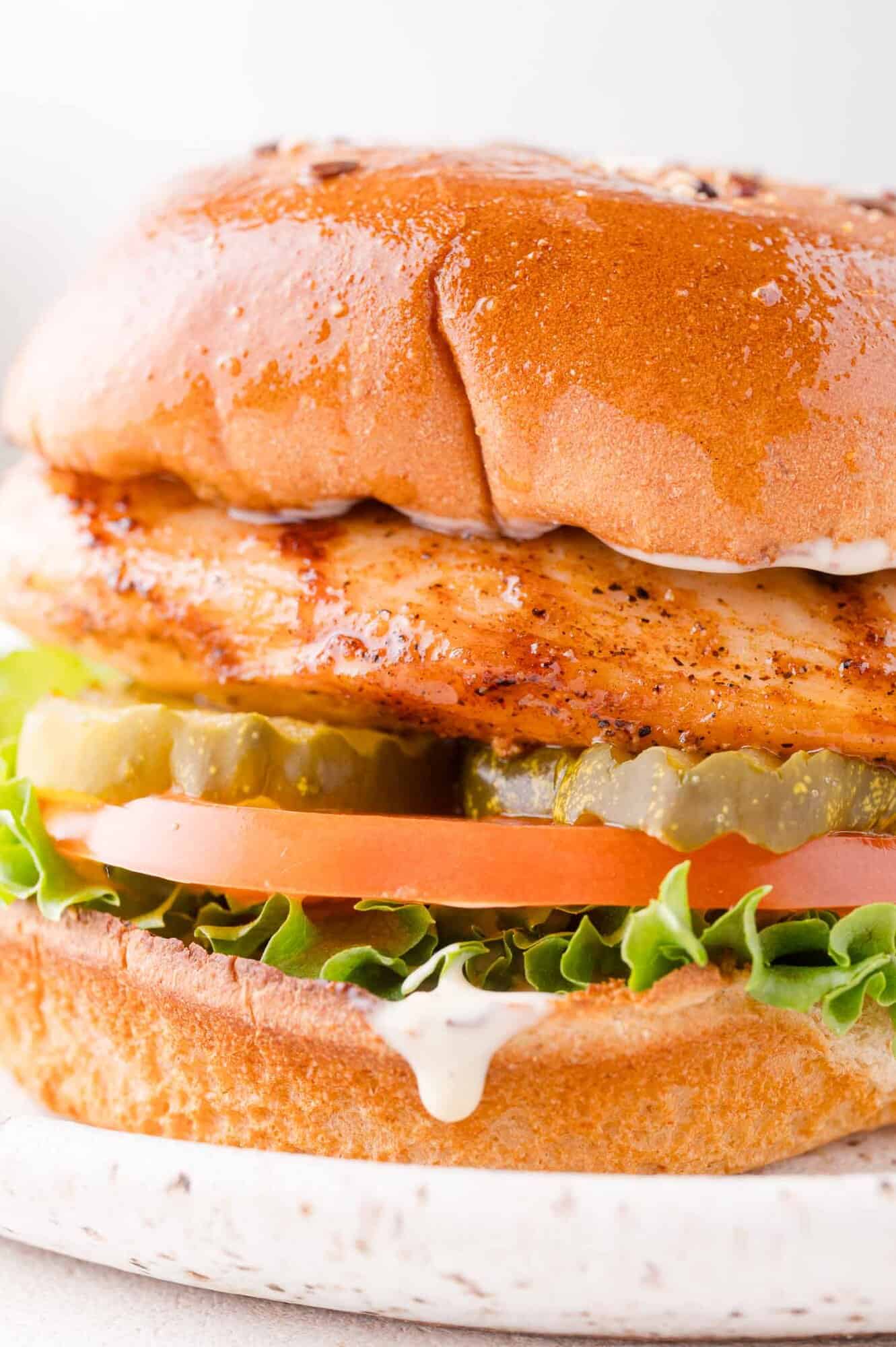Close-up of a grilled chicken sandwich with lettuce, tomato, pickles, and mayonnaise on a toasted bun.