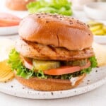 Grilled chicken sandwich with lettuce, tomato, pickles, and sauce on a seeded bun, served with potato chips on a white plate.