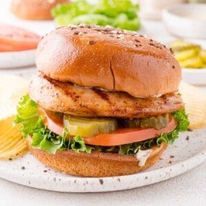 Grilled chicken sandwich with lettuce, tomato, pickles, and sauce on a seeded bun, served with potato chips on a white plate.