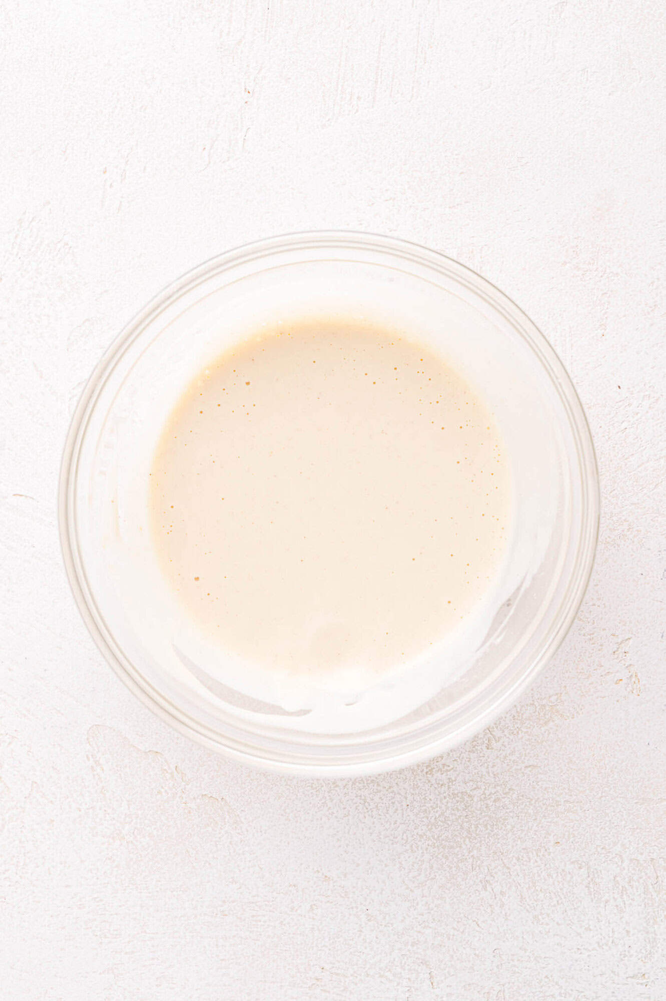 A clear glass bowl filled with a creamy, light beige liquid mixture sits on a white textured surface.