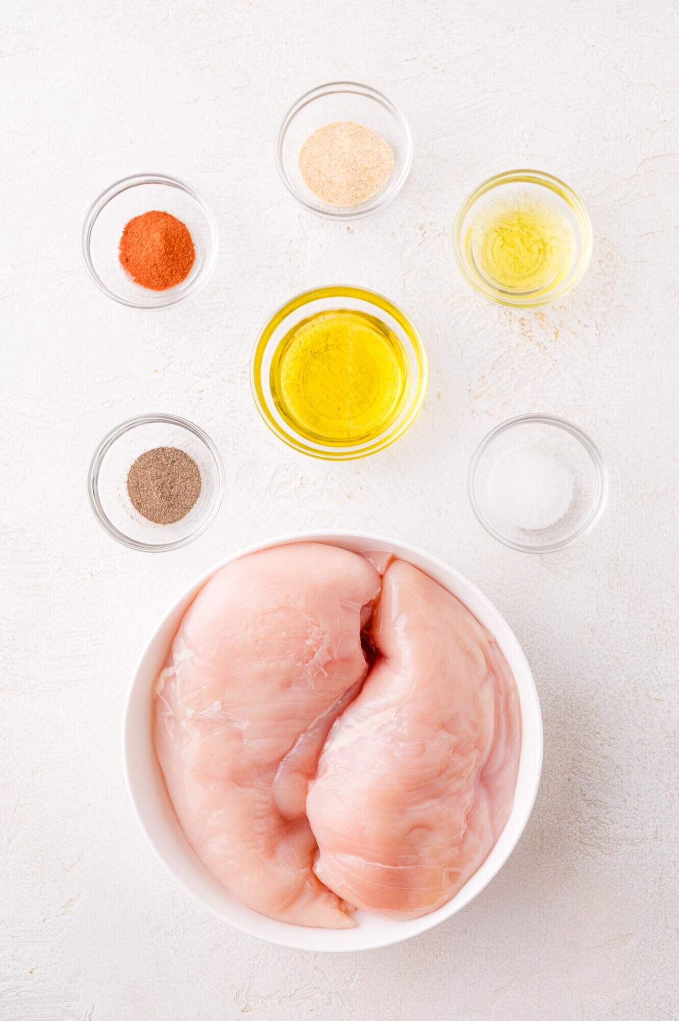 Raw chicken breasts in a white bowl surrounded by small glass bowls containing various spices, oil, and seasoning on a white surface.