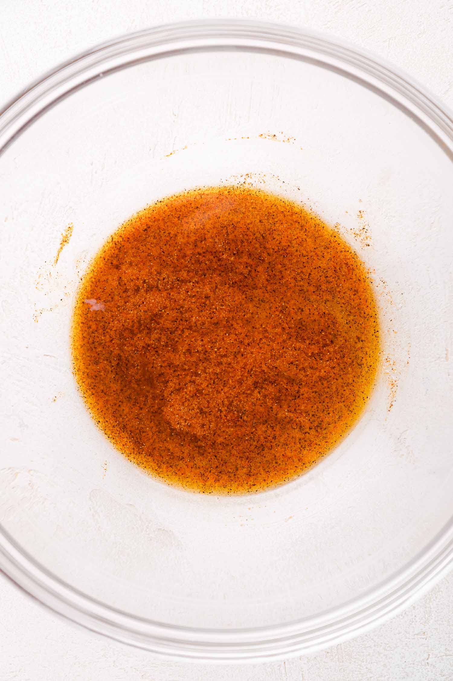A glass bowl containing a marinade - a mixture of brown liquid and specks of seasoning on a white surface.