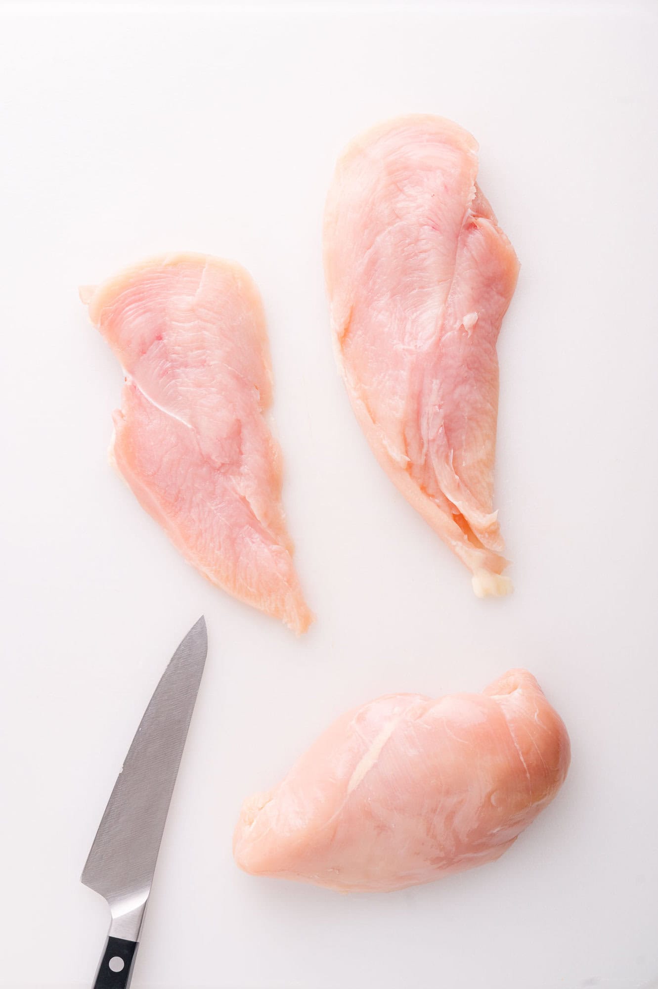 A raw chicken breast and two sliced chicken breast halves on a white cutting board with a kitchen knife nearby.