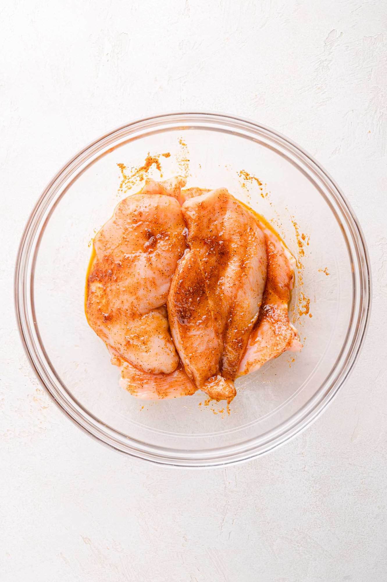 Two raw chicken breasts coated with seasoning in a clear glass bowl on a light-colored surface.