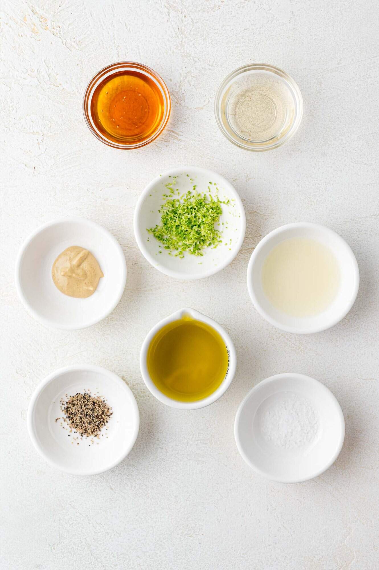 Seven small bowls containing various ingredients, including olive oil, black pepper, mustard, lime zest, lime juice, salt, honey, and vinegar, arranged on a white surface.