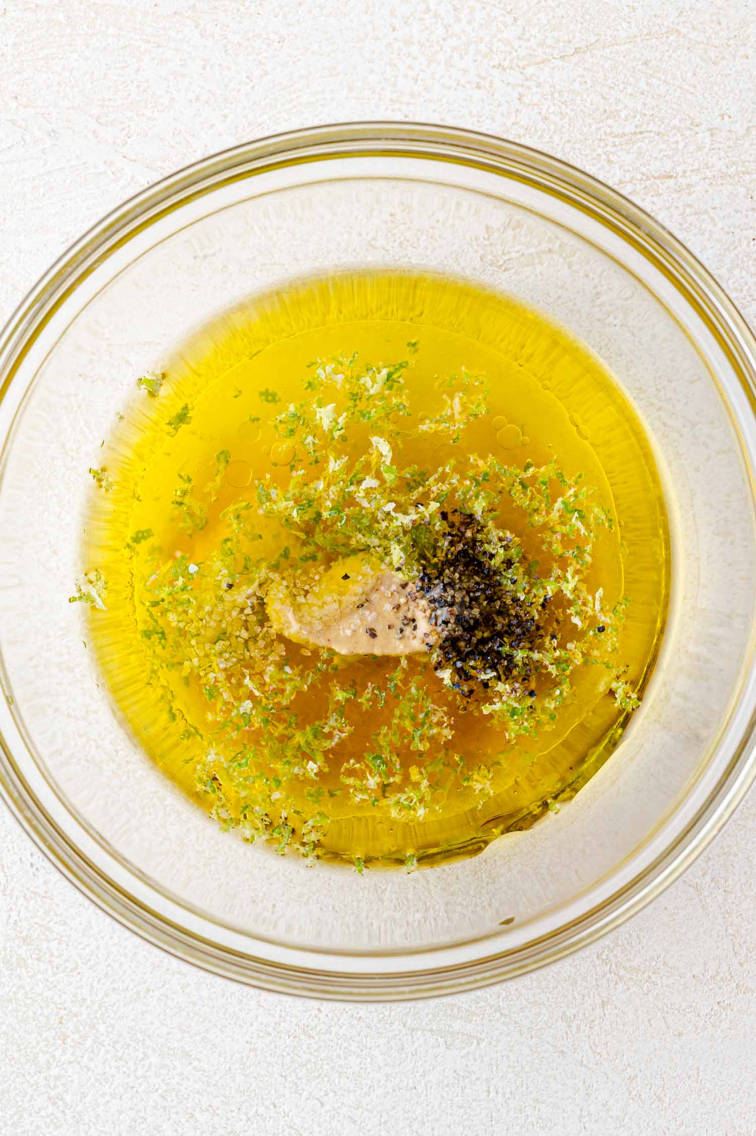 A glass bowl containing olive oil, mustard, black pepper, and lime zest on a light-colored surface.