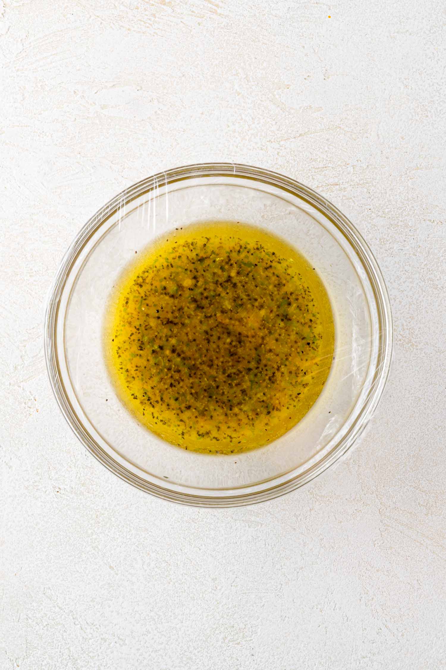 A glass bowl filled with yellow salad dressing mixed with herbs and spices, placed on a light textured surface.