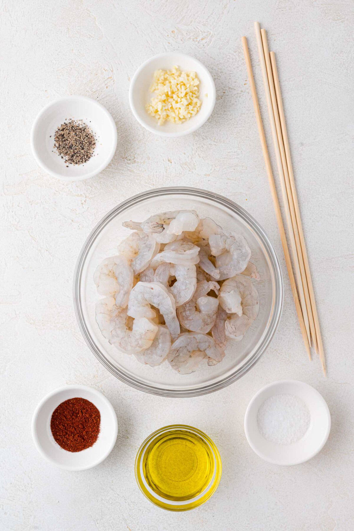 A bowl of raw shrimp surrounded by small bowls of minced garlic, black pepper, salt, paprika, and olive oil, with three wooden chopsticks on a white surface.